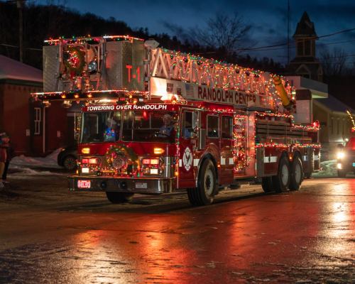 A decorated fire truck with lights drives through a snowy street at dusk, showcasing holiday spirit in Randolph Center.