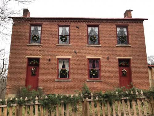 A brick house with red doors and windows decorated with wreaths, surrounded by greenery and a wooden fence.