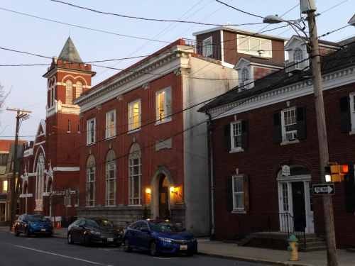 Historic red brick buildings line a quiet street at dusk, with cars parked along the curb and power lines overhead.