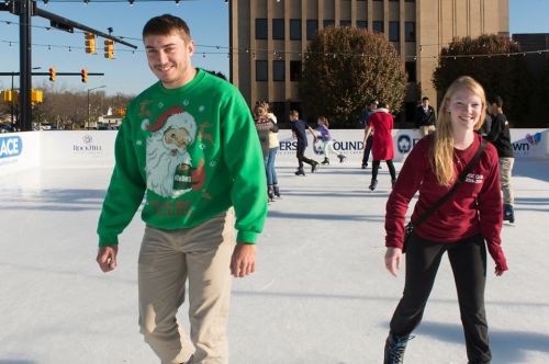 Two people ice skating outdoors, smiling. One wears a festive green sweater, the other a maroon sweatshirt.