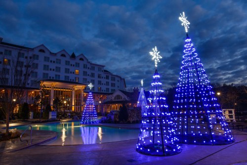 Illuminated Christmas trees with blue lights by a pool, set against a twilight sky and a hotel in the background.