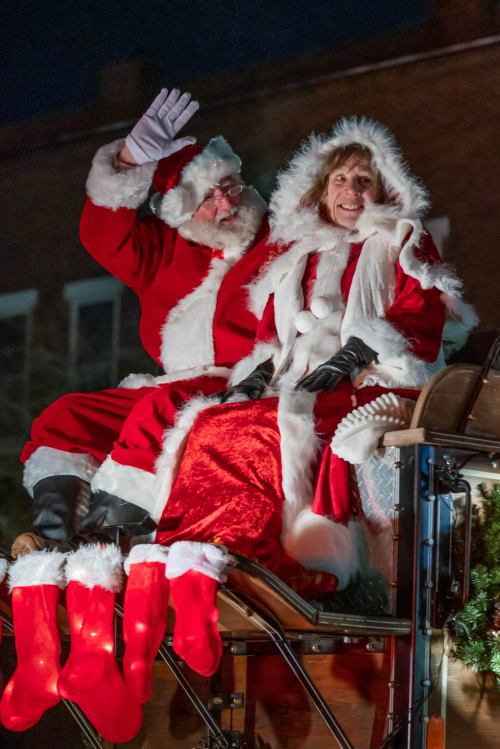 Santa Claus and a woman in festive attire wave from a decorated sleigh, surrounded by Christmas stockings.