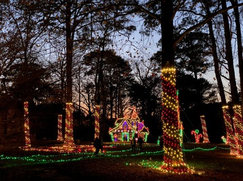 A festive scene with colorful Christmas lights on trees and a gingerbread house at dusk.