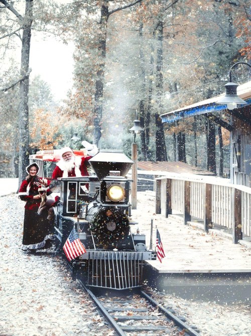 Santa and a woman in vintage attire wave from a festive train in a snowy, autumn forest setting.