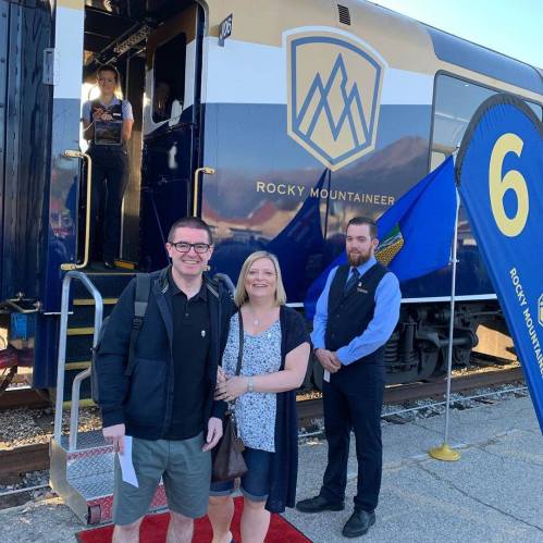 A couple poses in front of a Rocky Mountaineer train, with a staff member and train platform in the background.