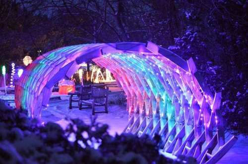 A colorful, illuminated archway in a snowy garden, with benches and festive lights in the background.