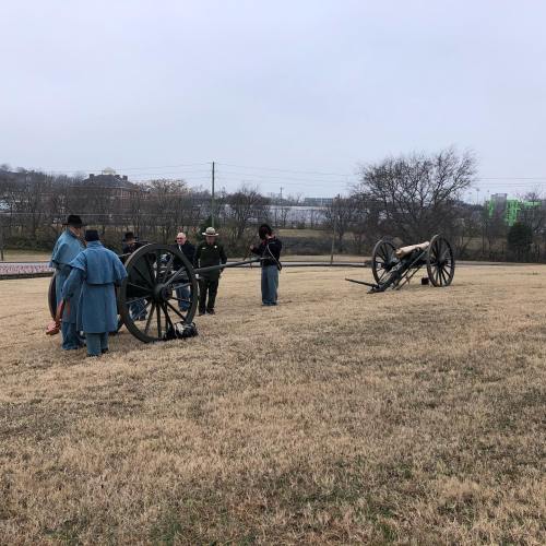 A group of people in historical attire stands near two cannons on a grassy field, with trees and buildings in the background.