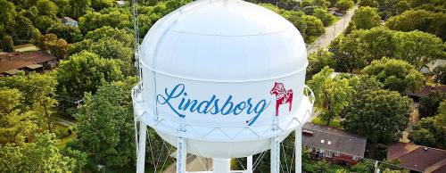 Aerial view of a white water tower with "Lindsborg" painted on it, surrounded by green trees and houses.