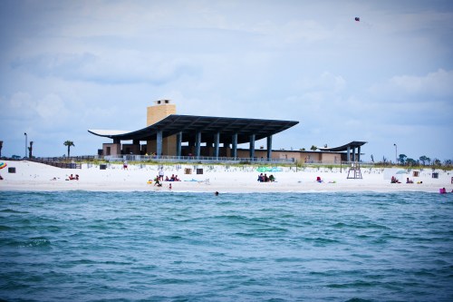 A beach scene featuring a modern building, white sand, and people enjoying the water under a cloudy sky.