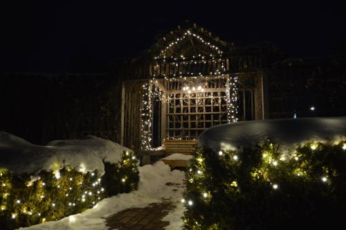 A snowy garden path leads to a lit gazebo adorned with string lights, creating a cozy winter atmosphere at night.