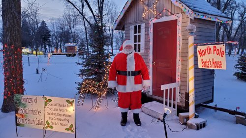 Santa Claus stands outside a decorated wooden cabin, surrounded by snow and festive lights, with signs for Santa's House.