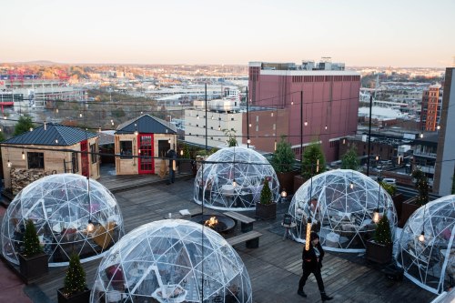 Rooftop view featuring transparent igloo-like structures, city skyline, and string lights at sunset.