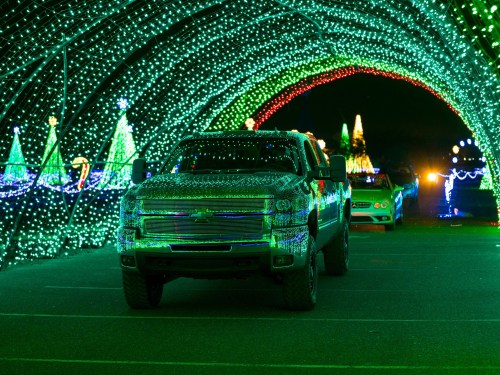 A truck covered in colorful lights drives through a tunnel of green and red holiday decorations.