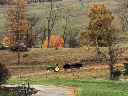 A green tractor pulls a wagon with people through a colorful autumn landscape with trees and rolling hills.