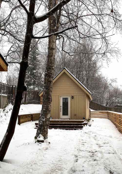 A small, beige cabin surrounded by snow, with a tree nearby and a wooden fence in the background.