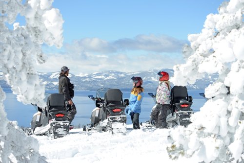 Three people in winter gear stand near snowmobiles, surrounded by snow-covered trees and mountains under a clear blue sky.