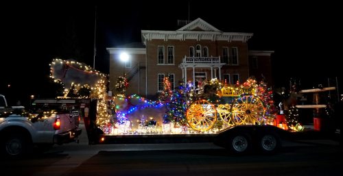 A festive parade float decorated with lights, trees, and a colorful wagon in front of a historic building at night.