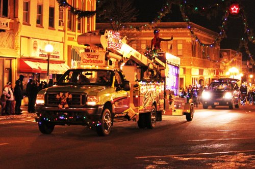 A festive parade float decorated with lights, featuring a person on top, driving through a lit-up street at night.