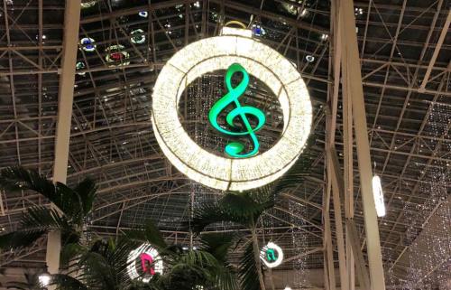 A large, illuminated musical note hangs from a ceiling in a festive indoor setting, surrounded by greenery.