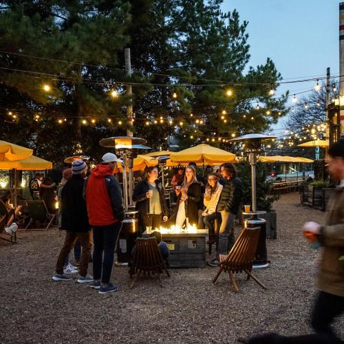 A group of people gathered around a fire pit in an outdoor setting, with string lights and patio umbrellas in the background.