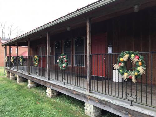 A wooden porch with festive wreaths hanging on the railing, surrounded by green grass and trees in the background.