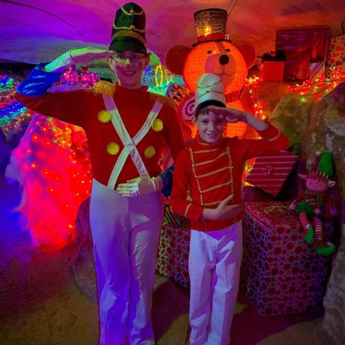 Two children in toy soldier costumes salute in a colorful, festive cave decorated with lights and holiday decorations.
