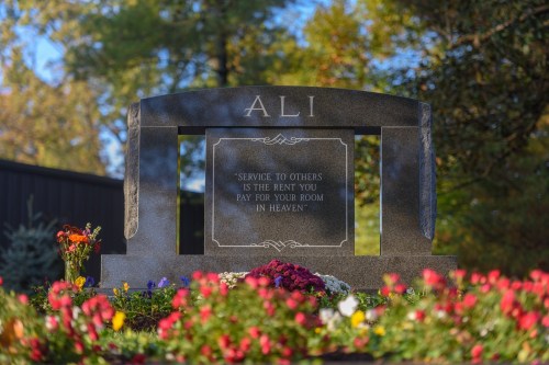 A gravestone with the name "ALI" and an inscription about service to others, surrounded by colorful flowers.