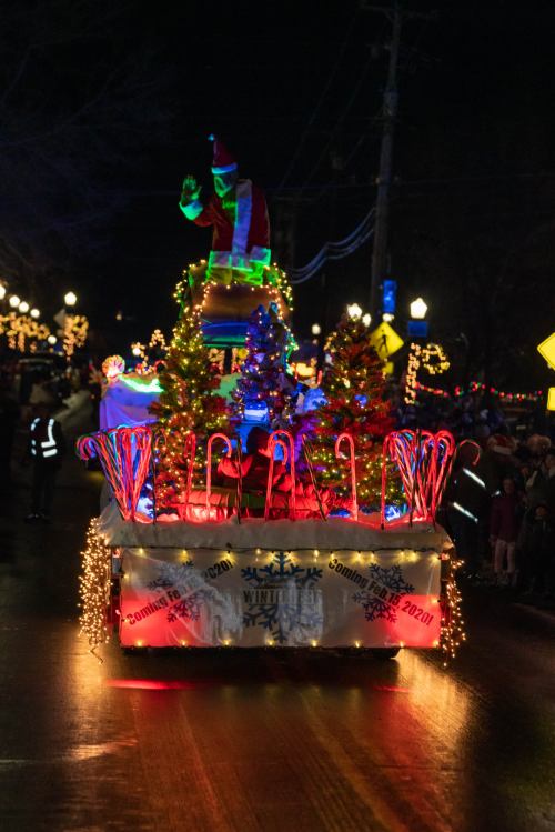 A festive parade float decorated with lights, featuring a Santa figure and colorful trees, moving through a nighttime street.