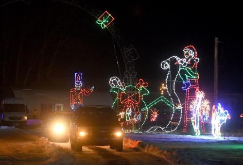 Colorful holiday lights display featuring Santa, a toy soldier, and festive decorations, with cars driving through snow.