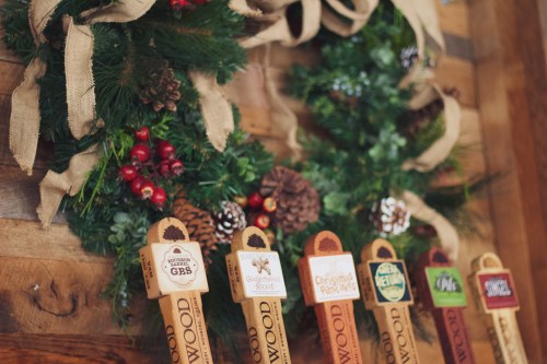 A festive display of beer tap handles arranged beneath a holiday wreath with pinecones and red berries.