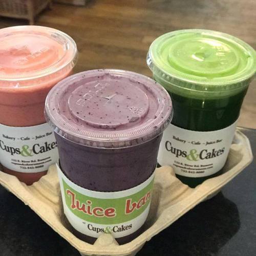 Three smoothie cups in a carrier: pink, purple, and green, labeled from "Cups & Cakes" and "Juice Bar."