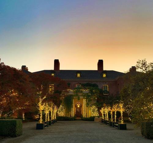 A grand mansion adorned with twinkling lights, surrounded by trees, at dusk with a colorful sky.