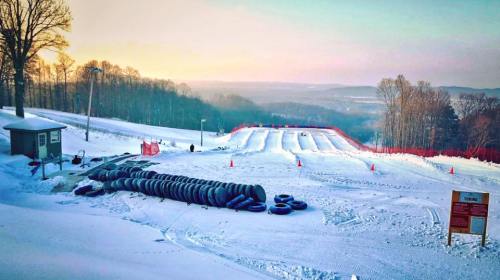 Snowy landscape featuring a tubing hill with stacked inner tubes, a small building, and trees in the background.