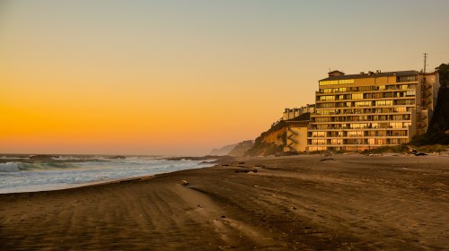 A beach at sunset with gentle waves and a modern building on the shore, surrounded by cliffs and a colorful sky.