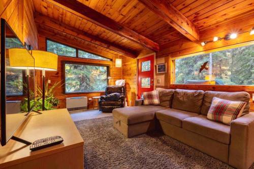 Cozy wooden cabin interior with a sectional sofa, TV, and large windows overlooking a forested view.