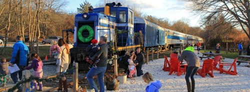 A blue train parked near a gathering of people, some taking photos, in a festive outdoor setting.
