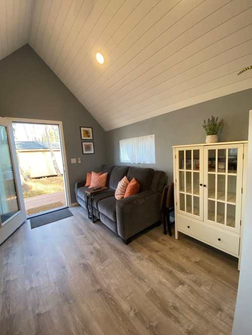Cozy interior of a room with a gray sofa, decorative pillows, a white cabinet, and large windows letting in natural light.