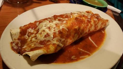 A large enchilada topped with melted cheese and red sauce, served on a white plate with a side of green sauce.