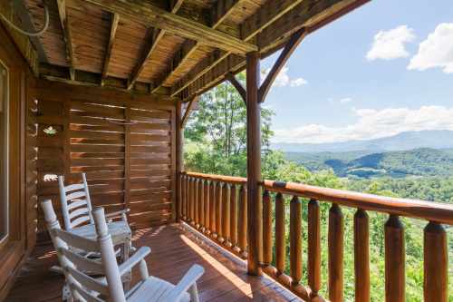 A wooden balcony with rocking chairs overlooks a lush green valley and distant mountains under a blue sky.