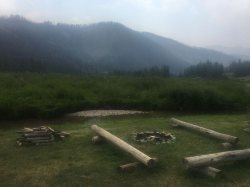 A serene landscape featuring a river, grassy area, and mountains in the background, with firewood and a fire pit nearby.