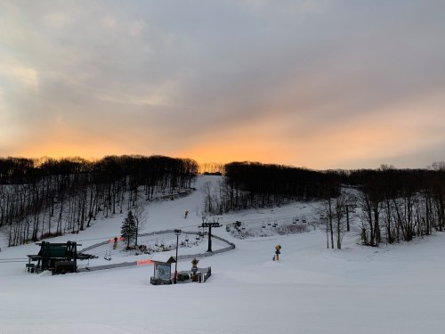 A snowy ski slope at sunset, with a ski lift and trees in the background.