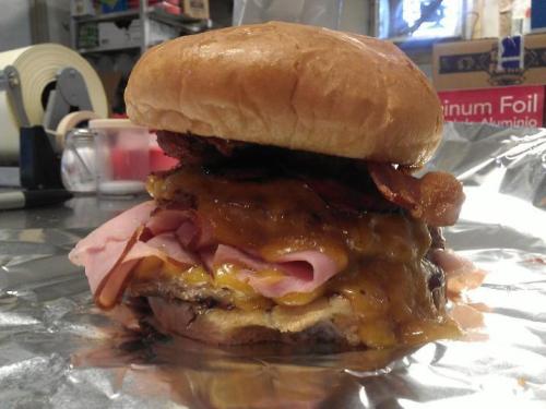 A towering burger with layers of cheese, ham, bacon, and a soft bun, resting on foil in a casual setting.