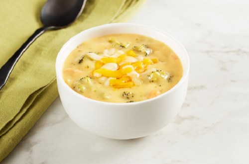 A bowl of creamy broccoli cheese soup topped with shredded cheese, placed on a marble surface next to a green napkin.