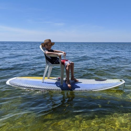 A child sits in a chair on a surfboard in calm water, wearing a hat and enjoying a sunny day.