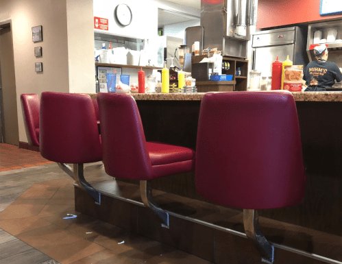 Red vinyl bar stools line a counter in a diner, with condiments and a kitchen visible in the background.