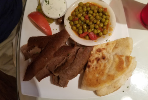 A plate with sliced meat, flatbread, green peas with tomatoes, olives, and a side of dipping sauce.