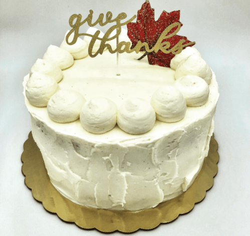 A white frosted cake with a "give thanks" topper and a red maple leaf, decorated with swirls of frosting around the edge.