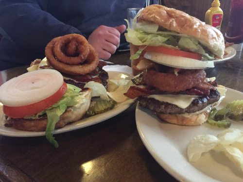 Two large burgers on plates, one topped with onion rings, lettuce, tomato, and pickles, served with potato chips.