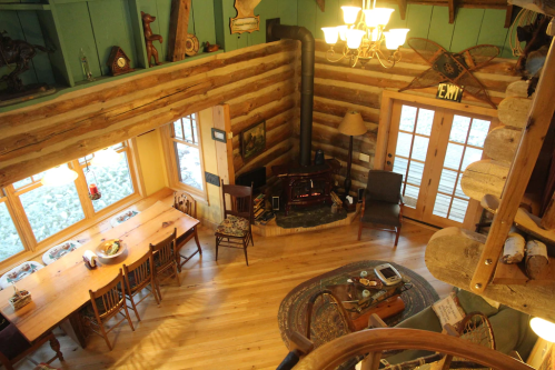 Cozy log cabin interior with a dining table, fireplace, and wooden decor, featuring large windows and a spiral staircase.