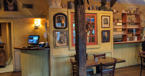 Interior of a cozy restaurant featuring a wooden counter, framed portraits on the walls, and a rustic table setting.
